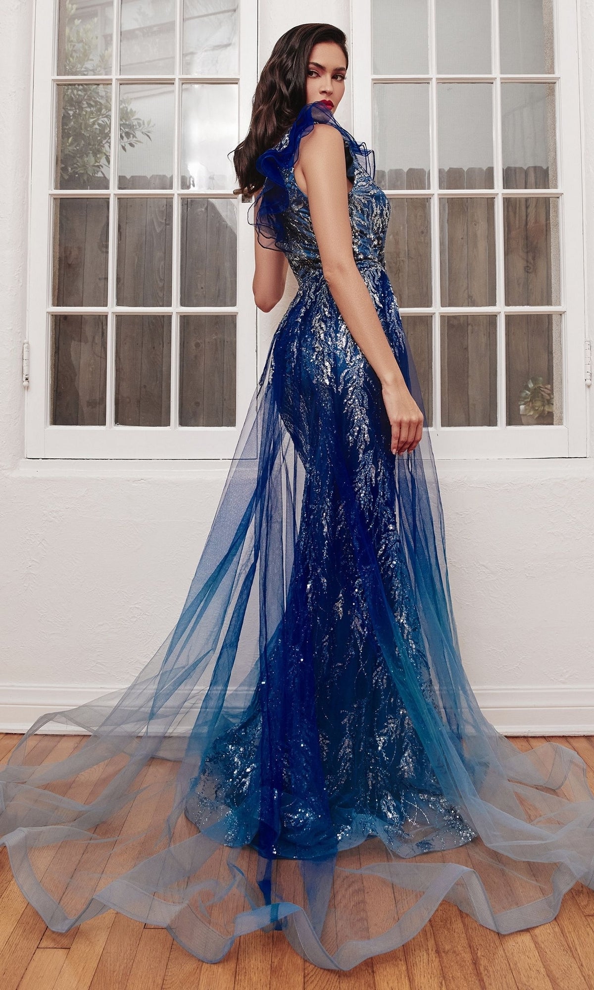 A woman poses indoors in front of large windows, wearing the Sheer-Overskirt Long Glitter Prom Dress J851, a floor-length blue and silver gown with a sheer tulle overlay.